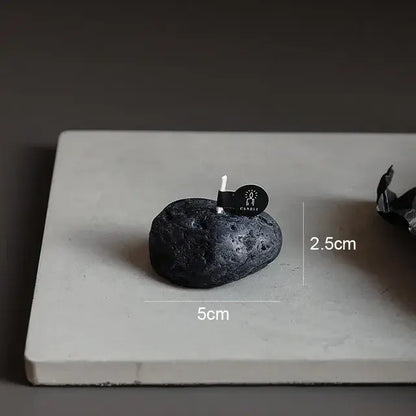 Bougie parfumée géométrique météorite noire sur surface blanche avec dimensions indiquées de 5 cm de largeur et 2,5 cm de hauteur.